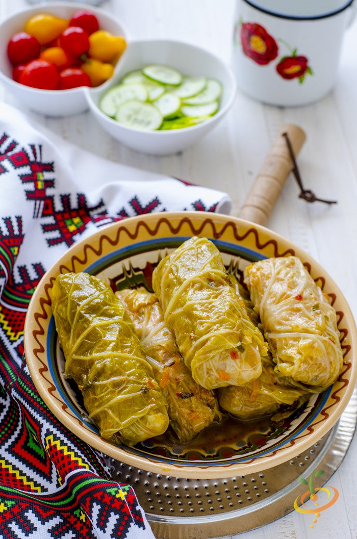 Plate of cabbage rolls made with All Season seeds cabbage leaves on a patterned cloth next to bowls of sliced cucumbers and cherry tomatoes
